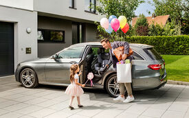 Vor einem Einfamilienhaus mit Vorgarten parkt ein graues Auto vor dem Garagentor. Ein Mann mit Tasche und Luftballone in der Hand öffnet einem kleinen Mädchen die Autotür. Auf der Rückbank im Auto ist ein Kindersitz montiert.