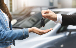 Eine Hand hält einen Autoschlüssel und gibt ihn einer anderen Person weiter. Im Hintergrund stehen zwei Autos.