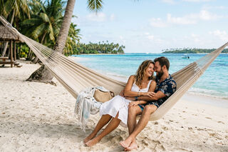 zeigt ein sich im Arm haltendes Paar mittleren Alters auf einer Hängematte an einem Palmenstrand