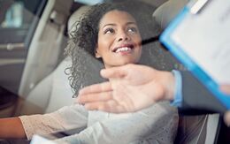 Lächelnde, junge Frau sitzt in einem Auto auf dem Fahrersitz während ein Mann mit einem Klemmbrett ihr etwas erklärt.