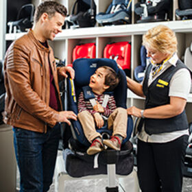 Ein Kleinkind sitzt in einem Kindersitz in einem Geschäft, links steht eine männliche Person und rechts eine Verkäuferin.