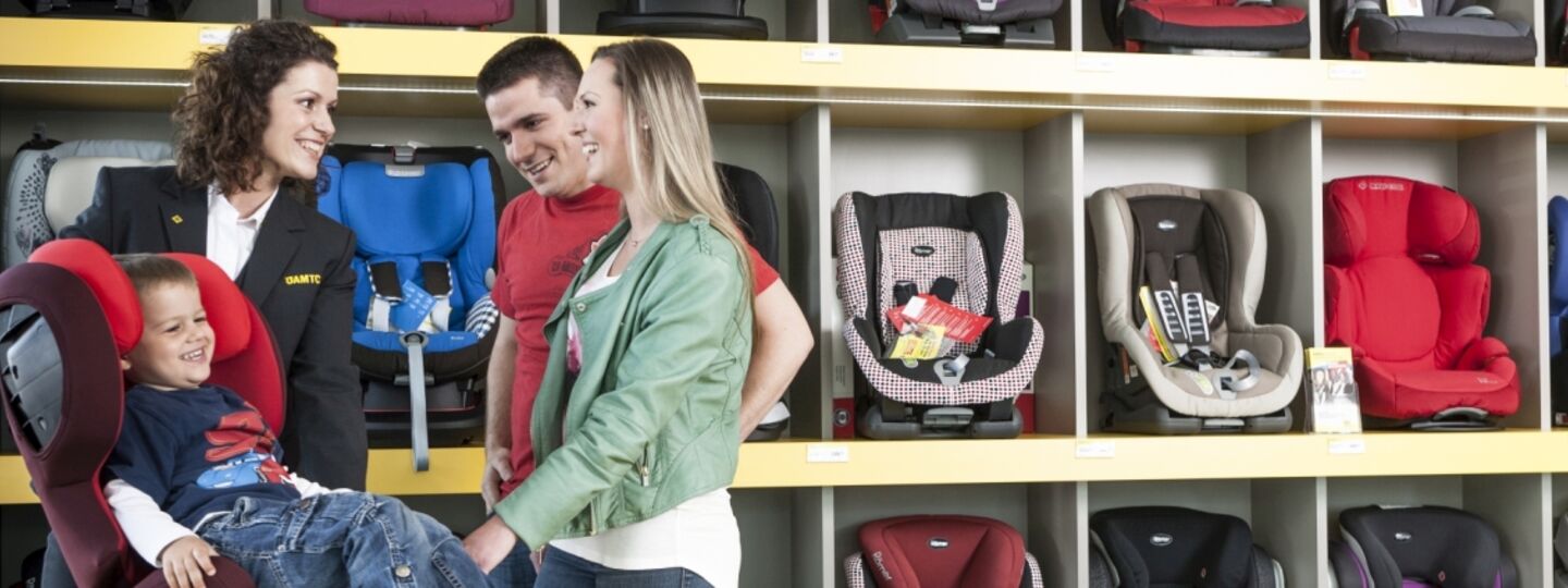 ÖAMTC-Mitarbeiterin zeigt Eltern mit Kind einen Kindersitz und erklärt die Sicherheitsmerkmale sowie die richtige Installation für den optimalen Schutz von Kindern im Auto.