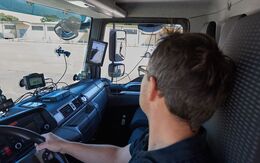 Lkw-Fahrer blickt nach rechts in den Seitenspiegel, wo ein Fußgänger sichtbar ist. Am Armaturenbrett befinden sich verschiedene Assistenzsysteme.