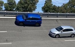 Ein blauer SUV und ein silberner Kleinwagen - Testautos für den Sommerreifentest 2022 - fahren hintereinander auf einer mehrspurigen Straße. Im Hintergrund sind Bäume und ein blauer Himmel zu sehen. 