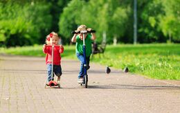 Ein kleines Mädchen und ein kleiner Junge fahren mit dem Tretroller bzw. Micro-Scooter auf einem Gehsteig im Park.