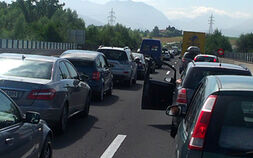 Stau auf einer Autobahn, viele Autos stehen auf beiden Fahrspuren in der Sonne. Im Hintergrund Landschaft mit Bäumen.